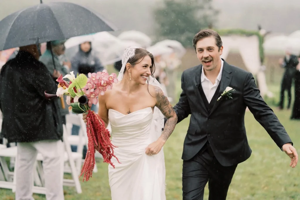 Newlyweds smiling as they walk back down the aisle after an outdoor wedding ceremony in the rain.