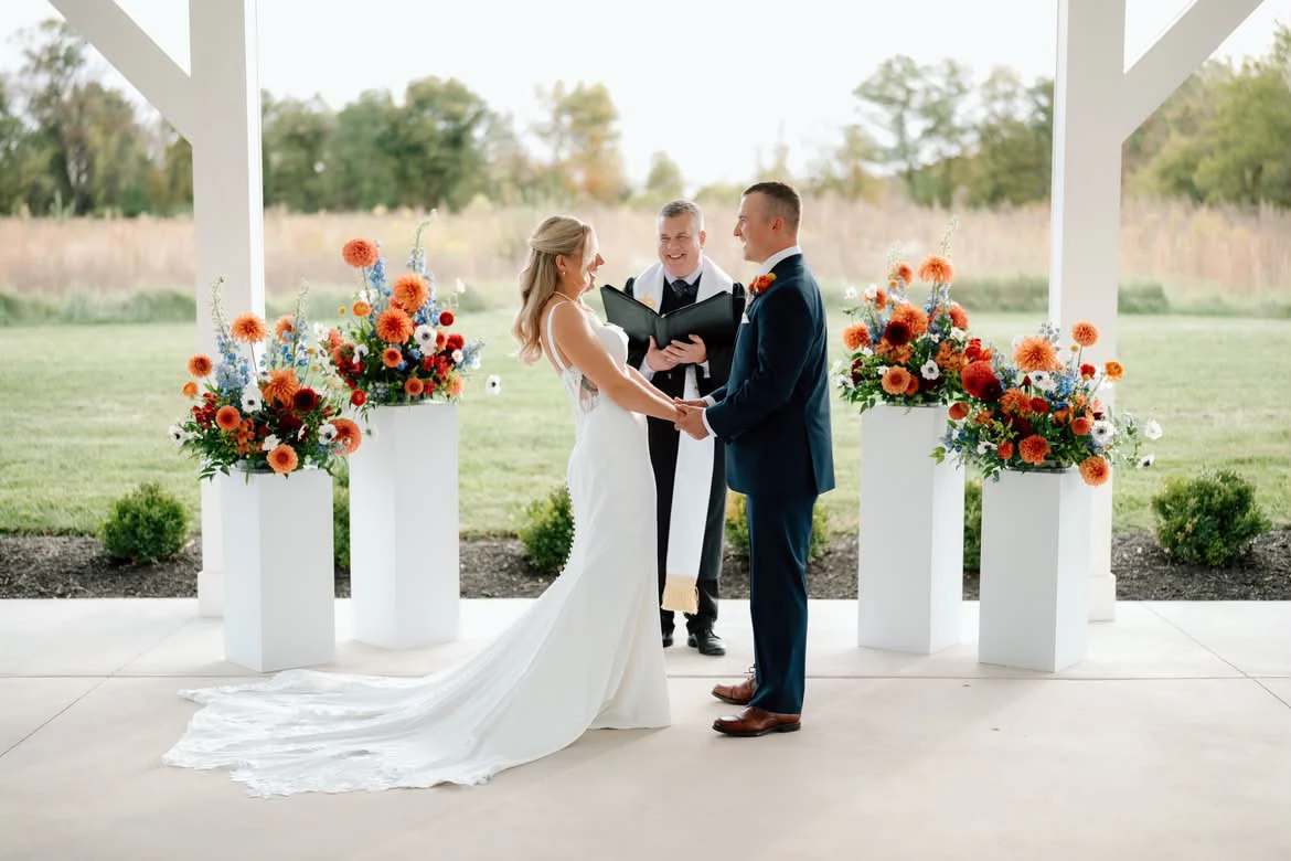 Kelly and Jordan at their outdoor autumn wedding ceremony, with Ever After Vows officiating beneath a pavilion framed by orange and blue flowers.