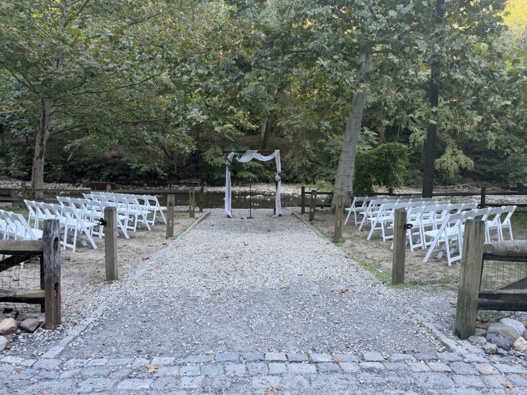 A gravel aisle between rows of white wedding chairs leads to a flower-draped arbor beside Wissahickon Creek in Philadelphia.