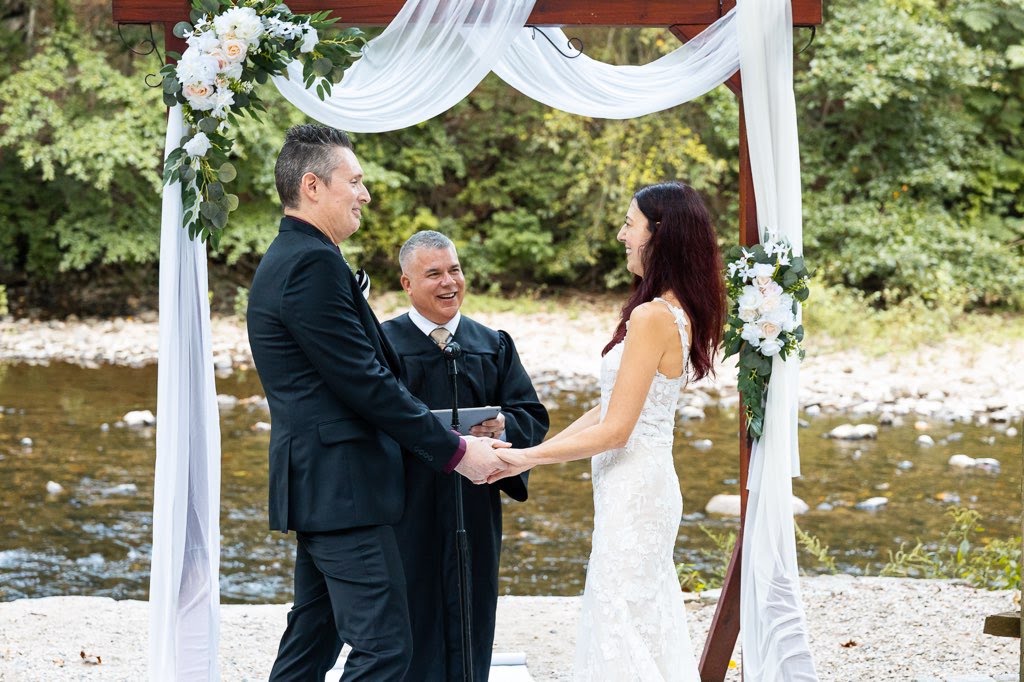 Sabrina and Nate hold hands beneath a flower-draped arbor beside Wissahickon Creek as officiant France Krazalkovich leads their wedding ceremony.