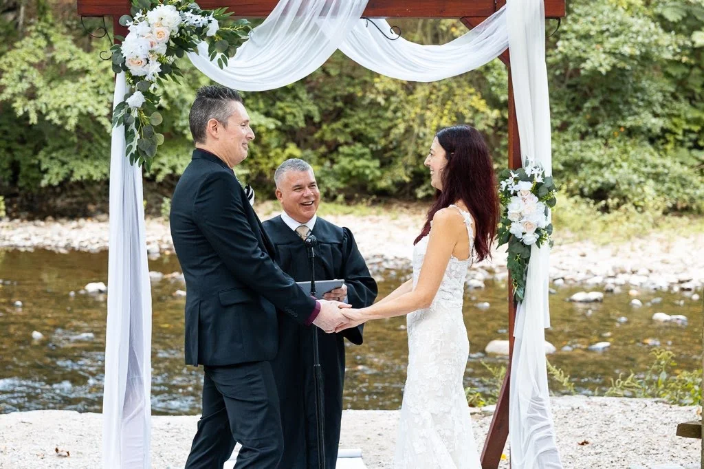 Sabrina and Nate hold hands beneath a flower-draped arbor beside Wissahickon Creek as officiant France Krazalkovich leads their wedding ceremony.