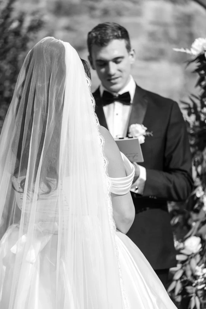 Groom reads from a vow booklet while facing his bride in a veil during an intimate outdoor wedding ceremony.